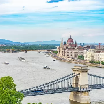 A river and a bridge above it connects to Budapest, Hungary. Historic buildings and a walking promenade line the waterfront. 
