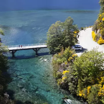 Two people on a narrow bridge over a waterway; a road is to one side of the bridge, curving around a cliff.