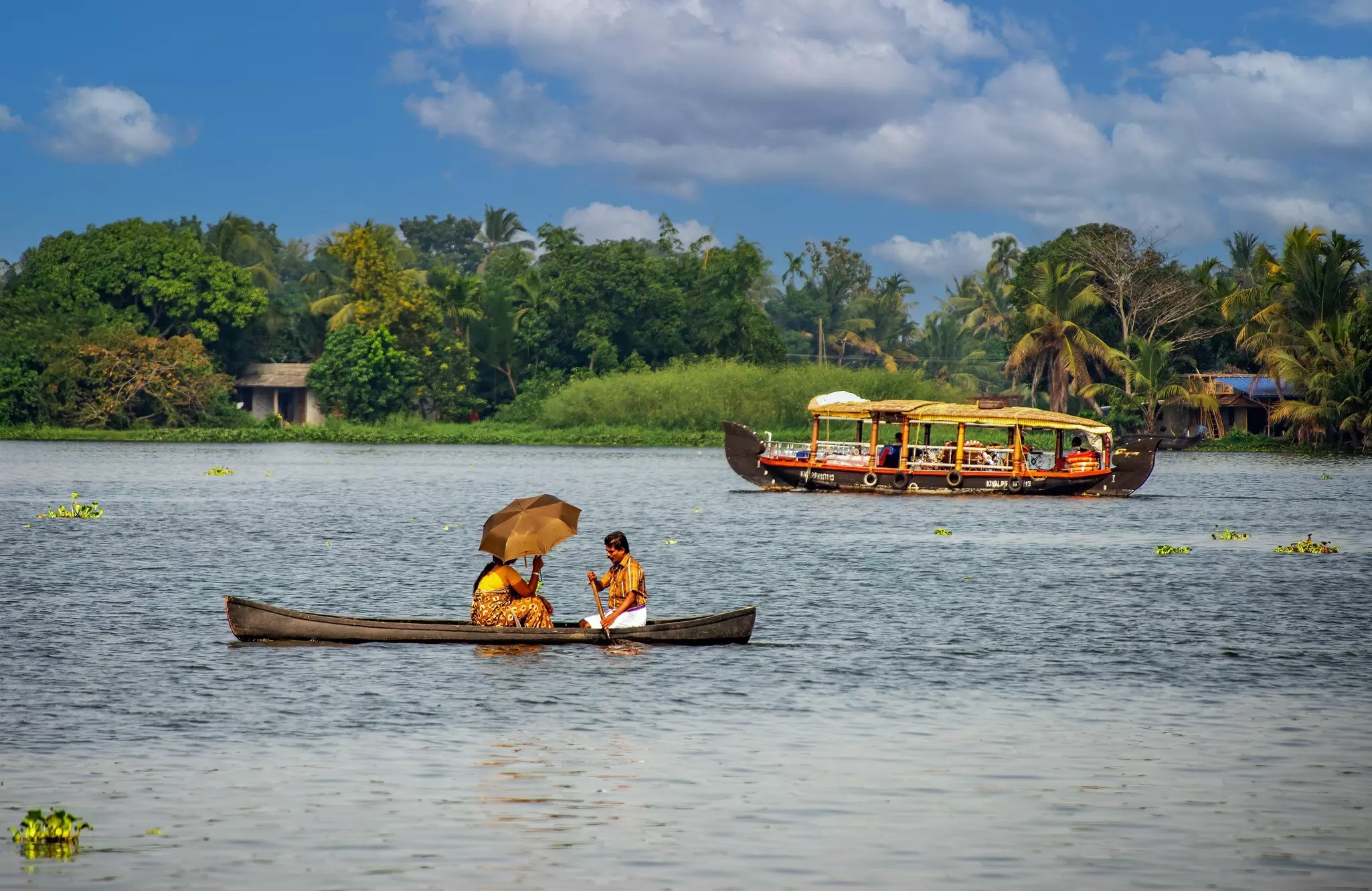 One person holds an umbrella and another rows a boat; a larger boat and a green shoreline are in the background.