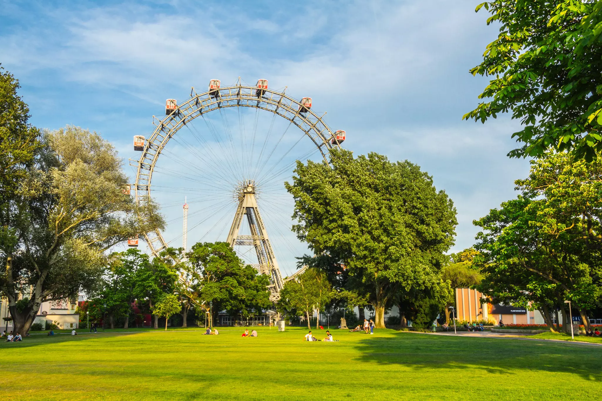 People relax on a large green lawn on a sunny day. A large Ferris wheel stands nearby