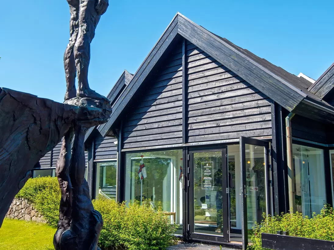 Tall bronze figure sculpture in front of the dark wooden gabled building of the Faroe Islands national art gallery