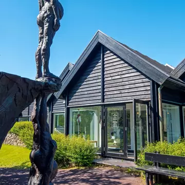Tall bronze figure sculpture in front of the dark wooden gabled building of the Faroe Islands national art gallery