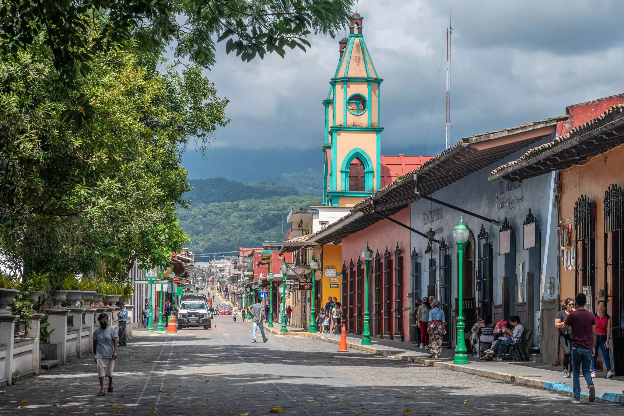 People walk along a cobblestone street on a sunny day, with colorful buildings to the right and forested mountains covered with clouds in the distance.
