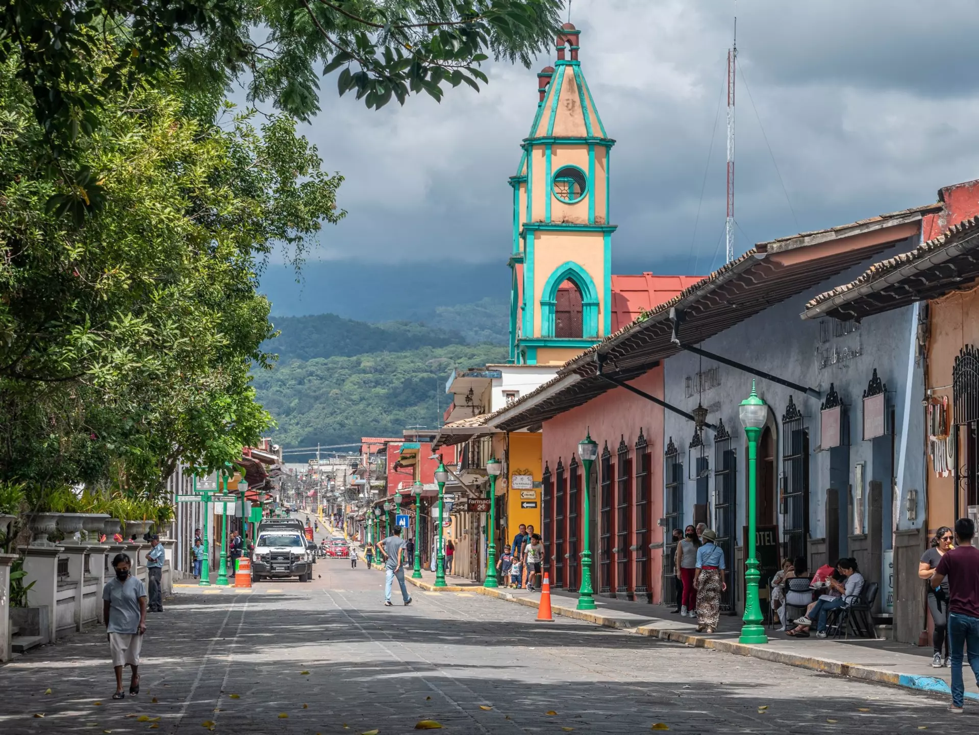 People walk along a cobblestone street on a sunny day, with colorful buildings to the right and forested mountains covered with clouds in the distance.
