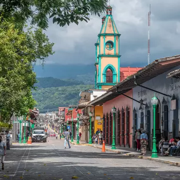 People walk along a cobblestone street on a sunny day, with colorful buildings to the right and forested mountains covered with clouds in the distance.