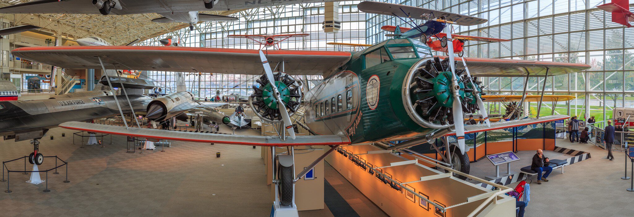 Green biplane with radial engines in a glass-walled hangar gallery; silver airliners and visitors behind it.