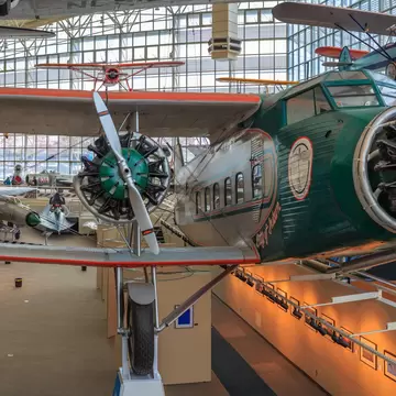 Green biplane with radial engines in a glass-walled hangar gallery; silver airliners and visitors behind it.