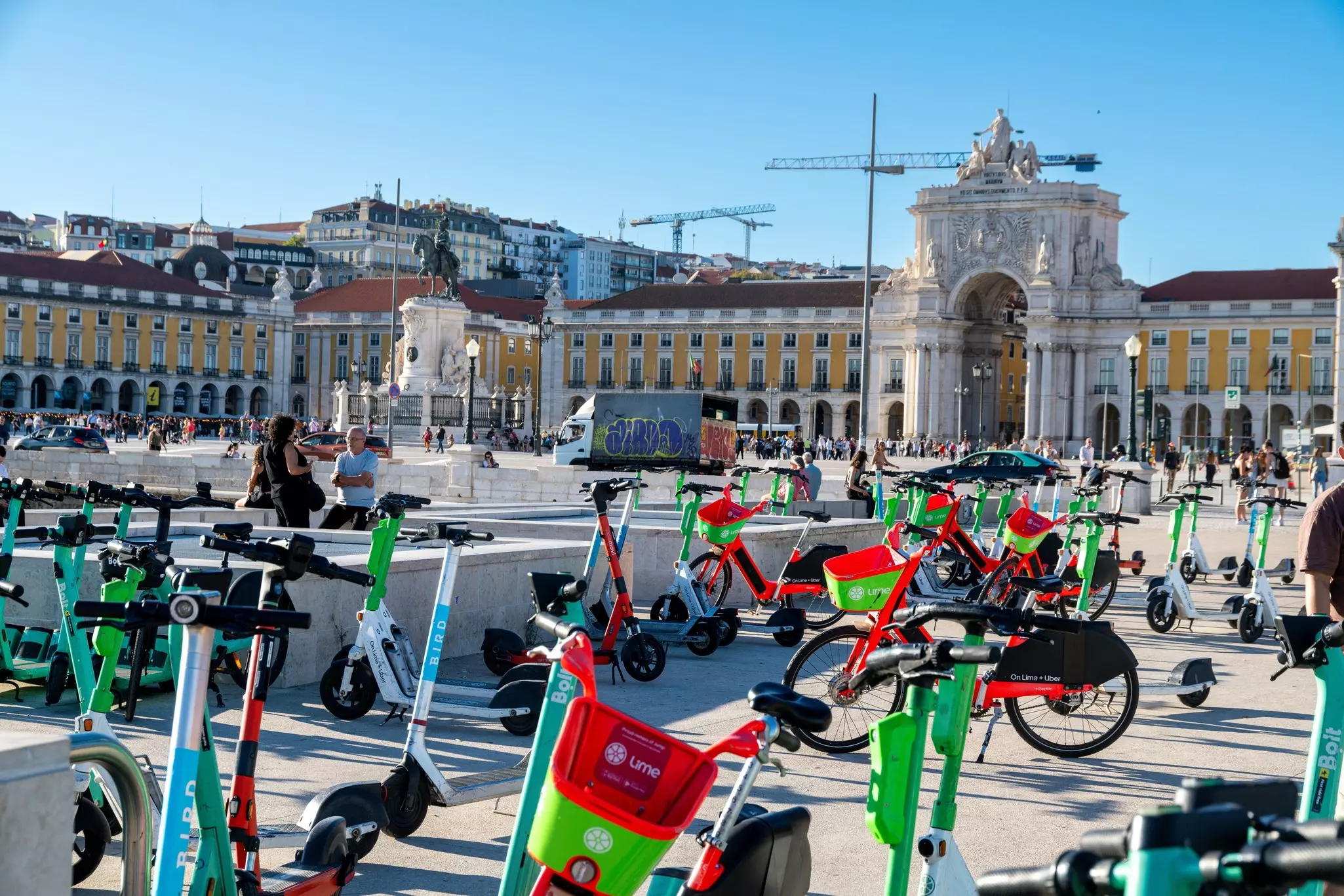 Colorful rental e-scooters and bikes in the foreground on  commercial plaza with statues and yellow buildings in the distance on a sunny day.