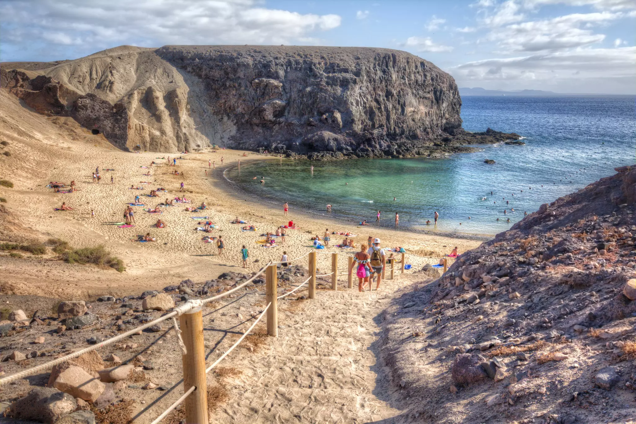 People descend steps to small beach with blue waters, in between jagged rock formations