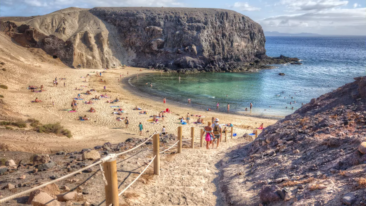 People descend steps to small beach with blue waters, in between jagged rock formations