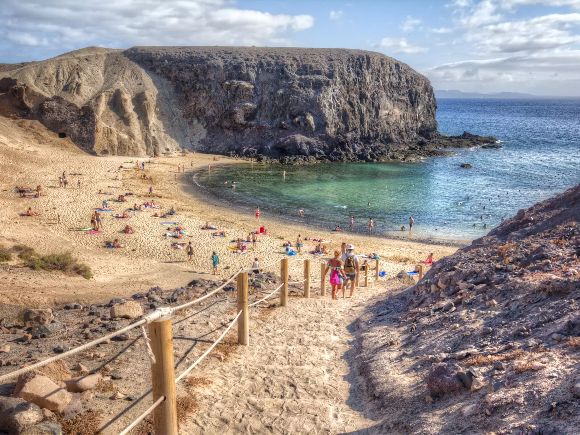 People descend steps to small beach with blue waters, in between jagged rock formations
