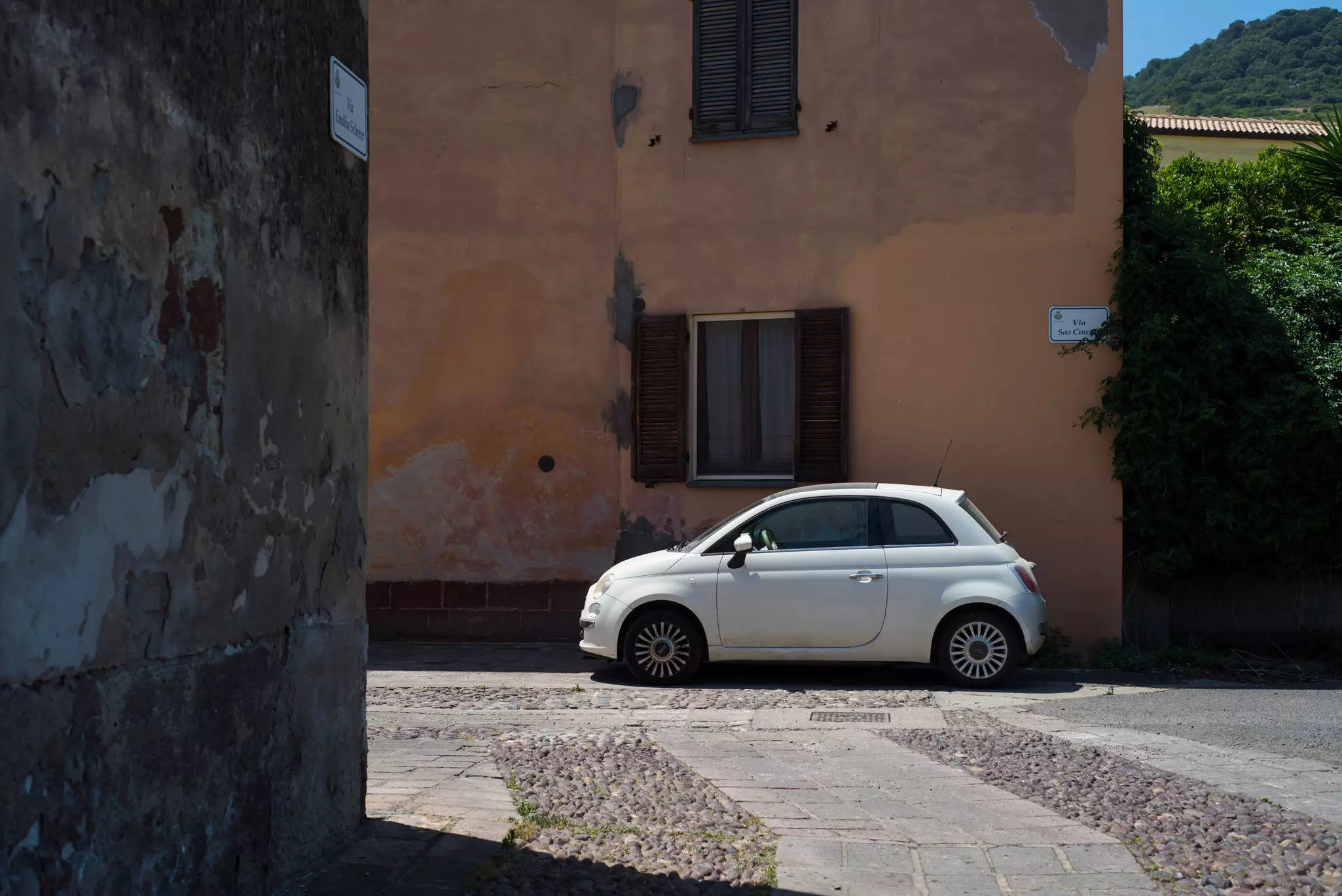 A compact white car is parked against a building in a narrow village street.