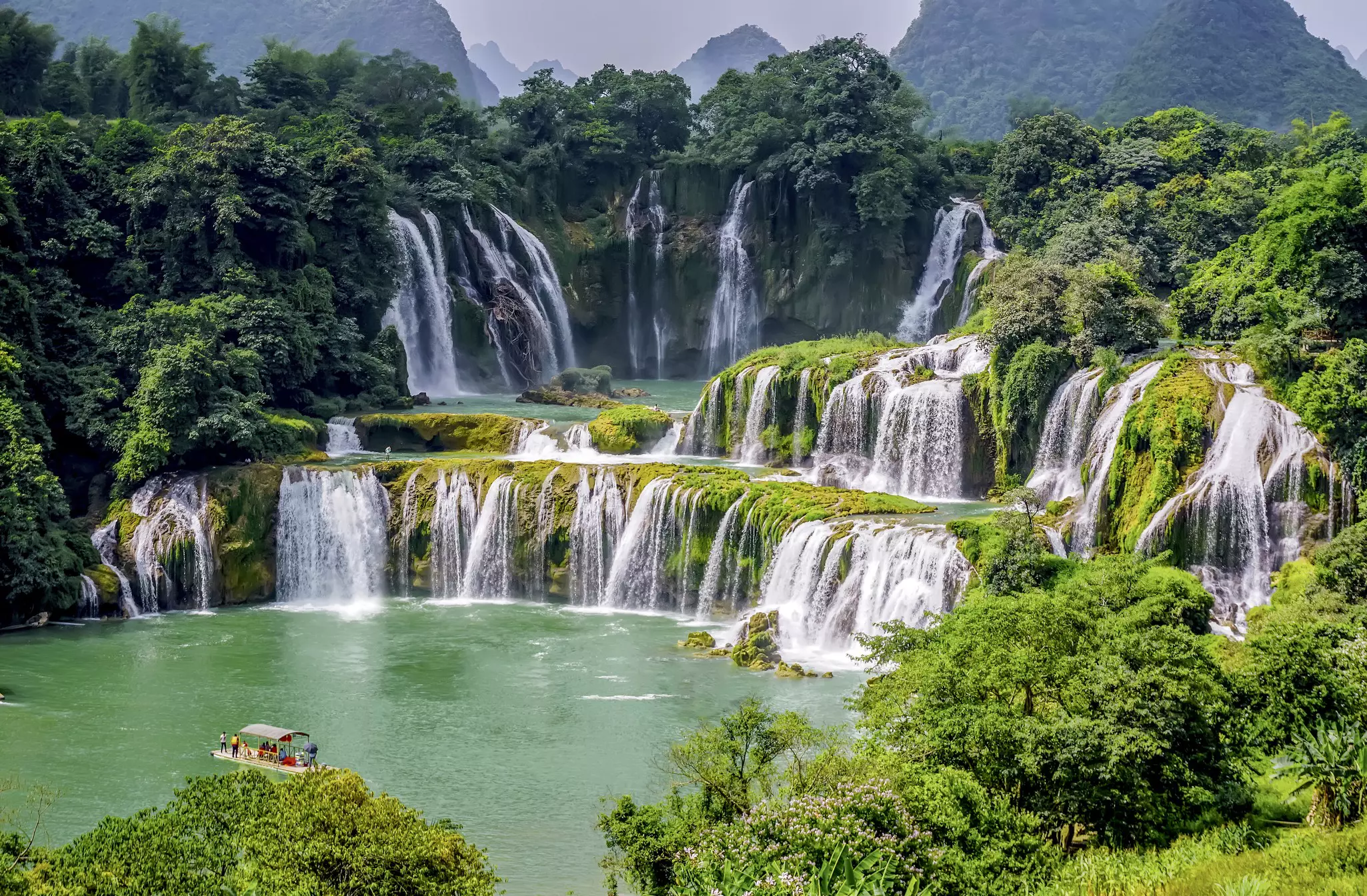 Several levels of waterfalls in Vietnam cascade out of a lush green landscape into a pool with a small viewing platform.