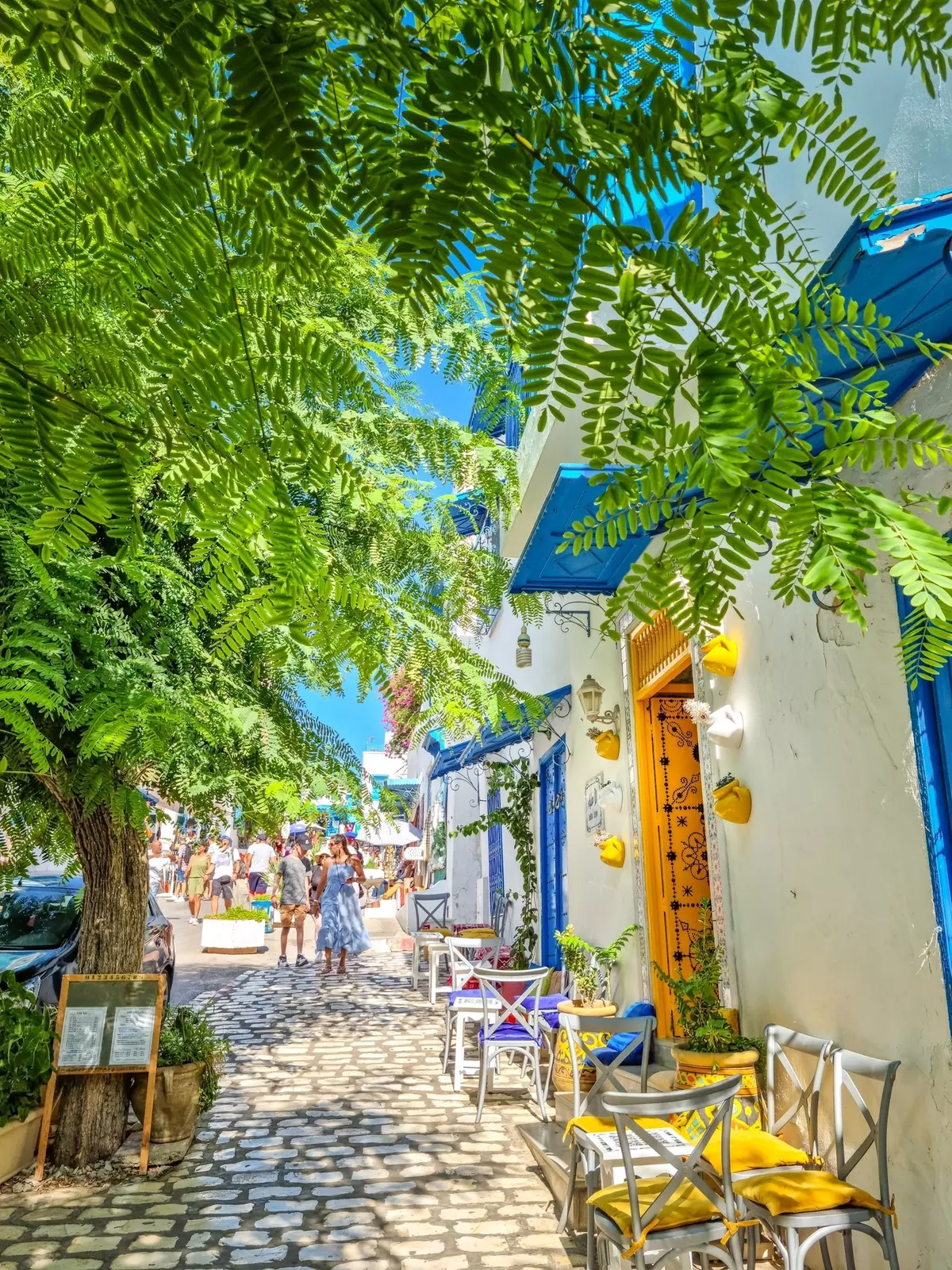 People walking down a cobblestone street with chairs set outside the buildings.