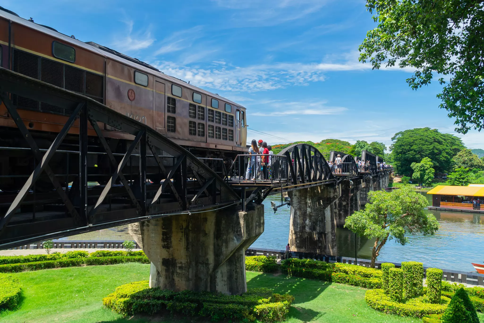 People stand on a steel train bridge across a river as an engine rolls by.