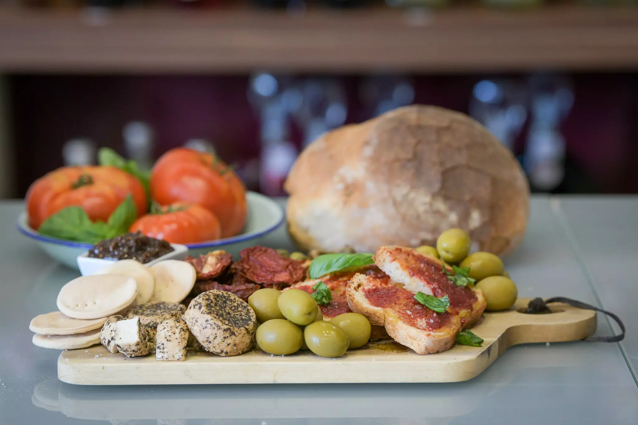 A wooden platter with olives, cheese, sundried tomatoes, crackers, sausage and a large round loaf of bread.