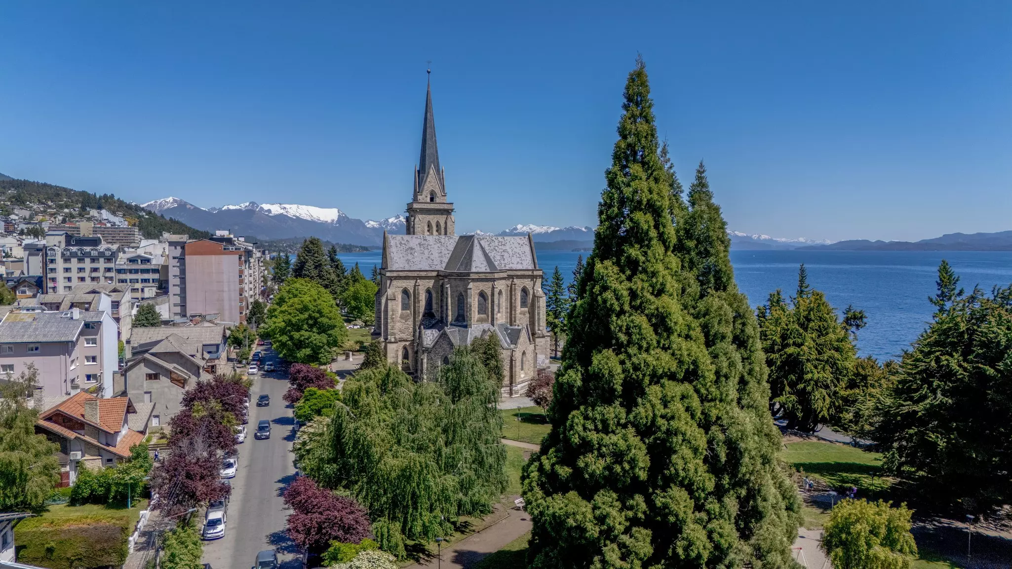 A town by a large blue lake has a large cathedral with a central spire; snowcapped mountains are in the distance.