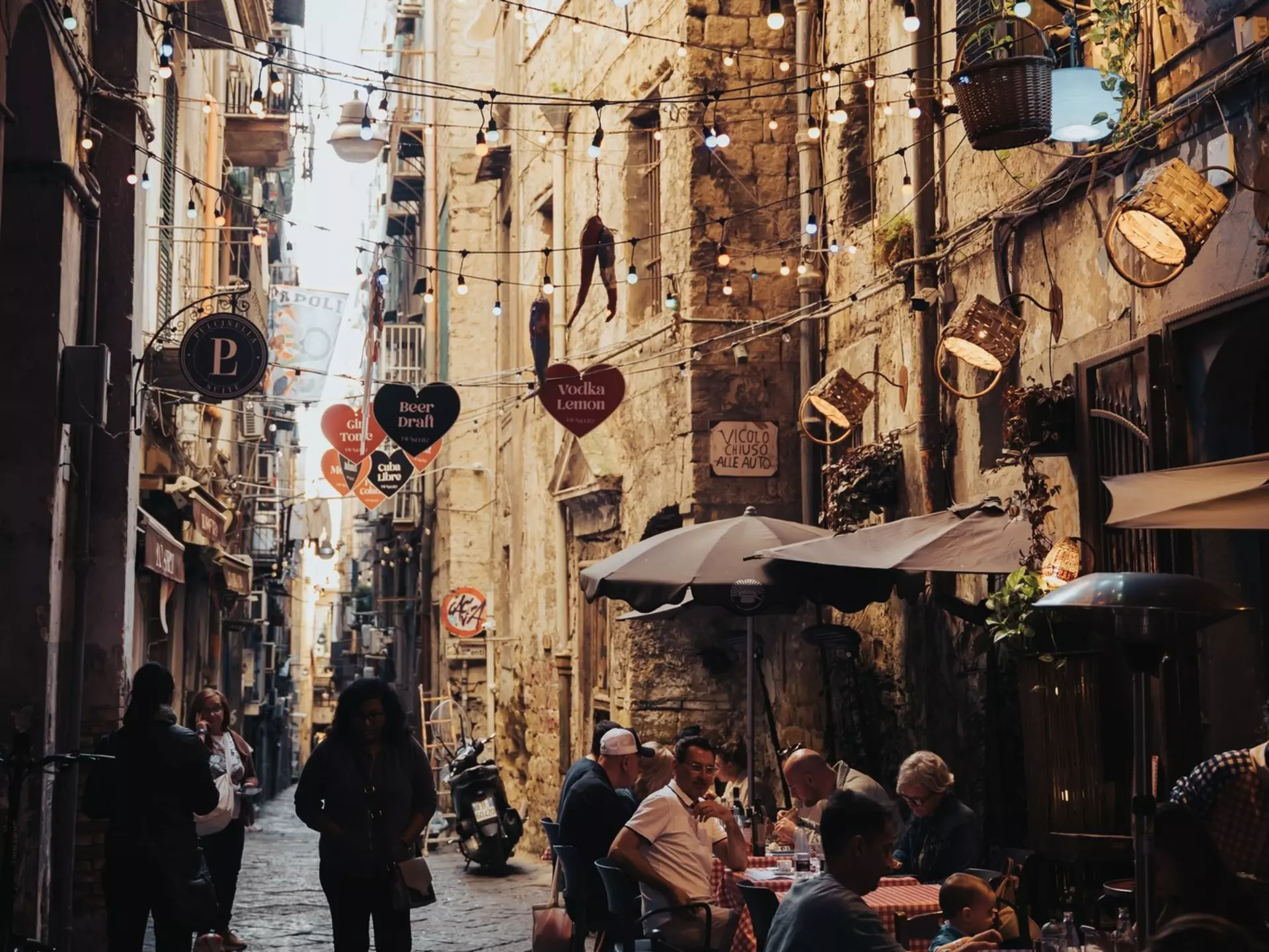 People sit at a sidewalk cafe in a shaded narrow alley.