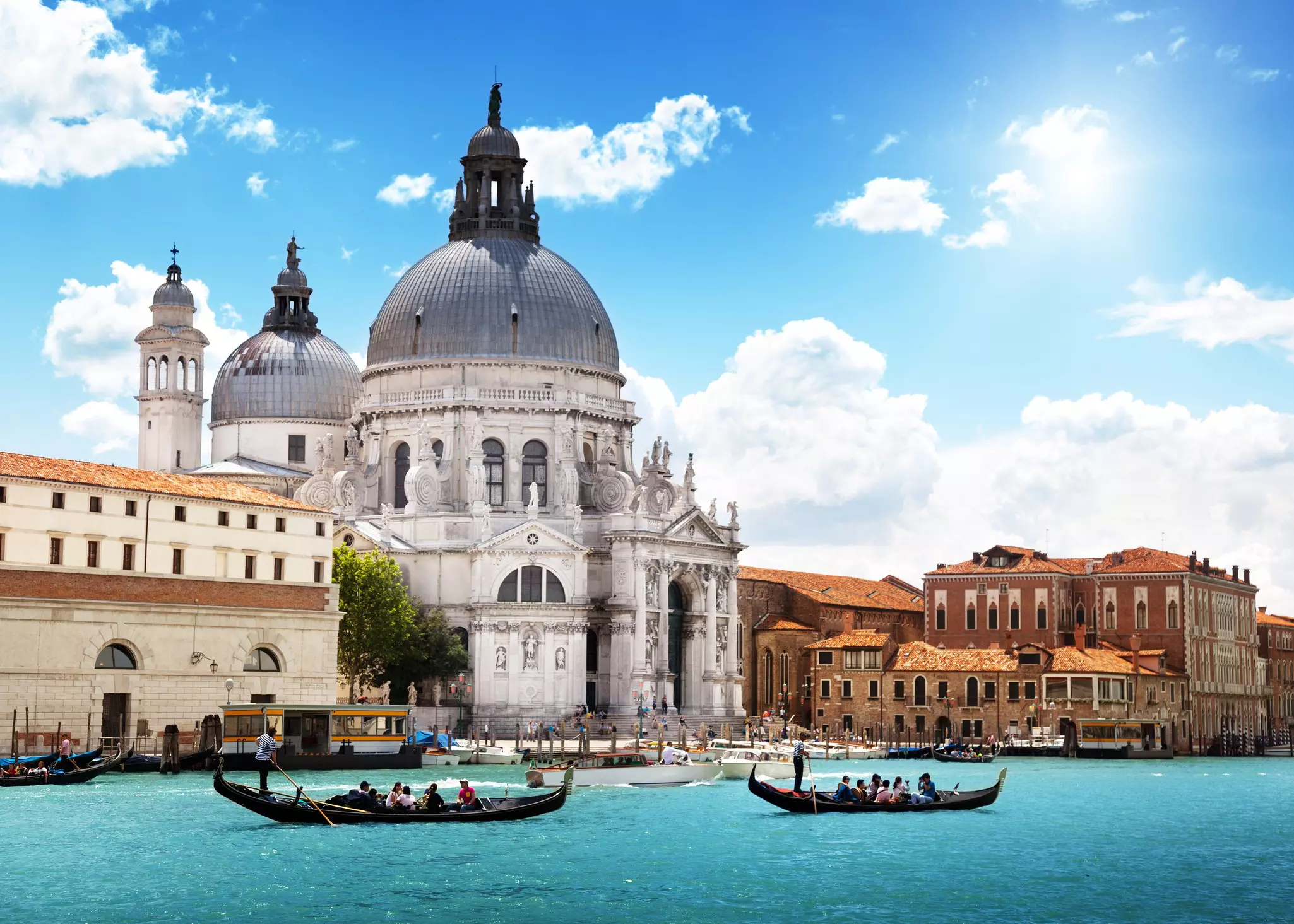 Two gondolas loaded with visitors pass by a grand domed church at the edge of a canal.