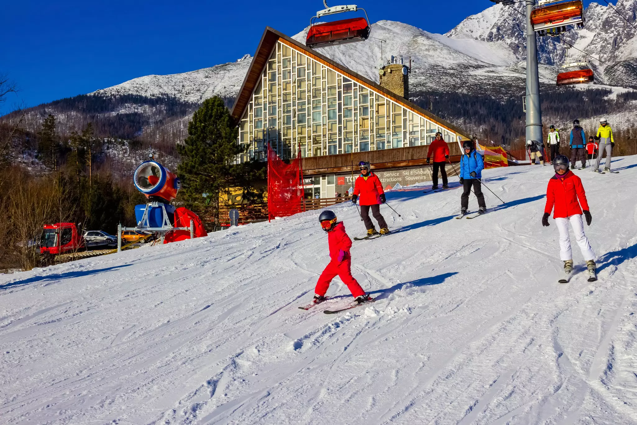 Skiers of many ages take to a slope at a mountain resort. A chalet-style lodge is seen in the distance.