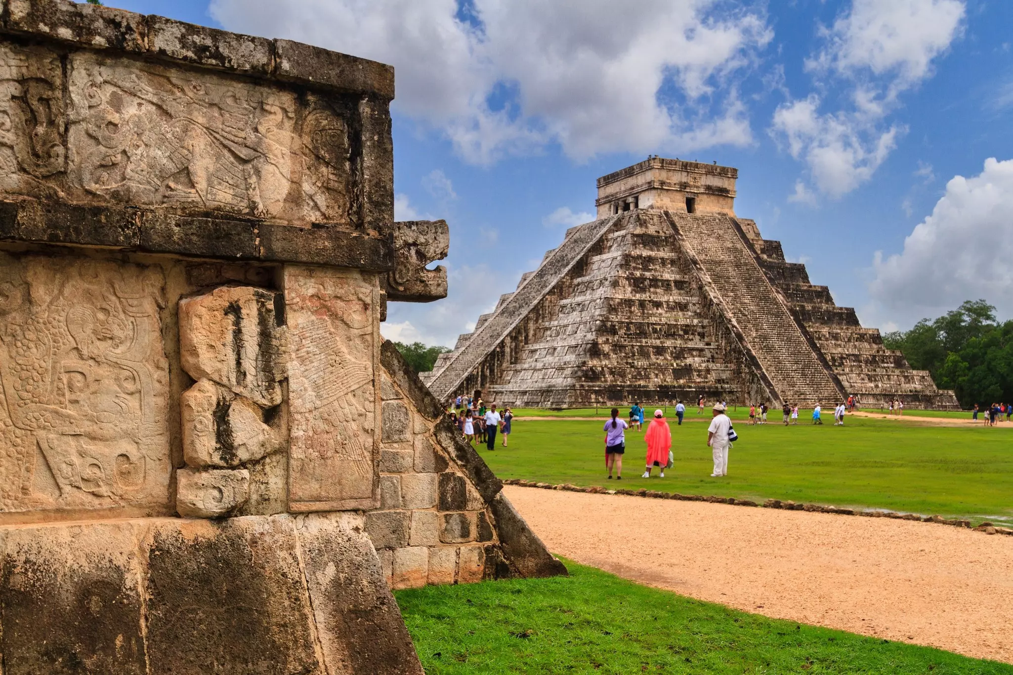 People gazing up at a vast stone pyramid viewed from a lawn. A nearby wall has ancient carvings in its brickwork