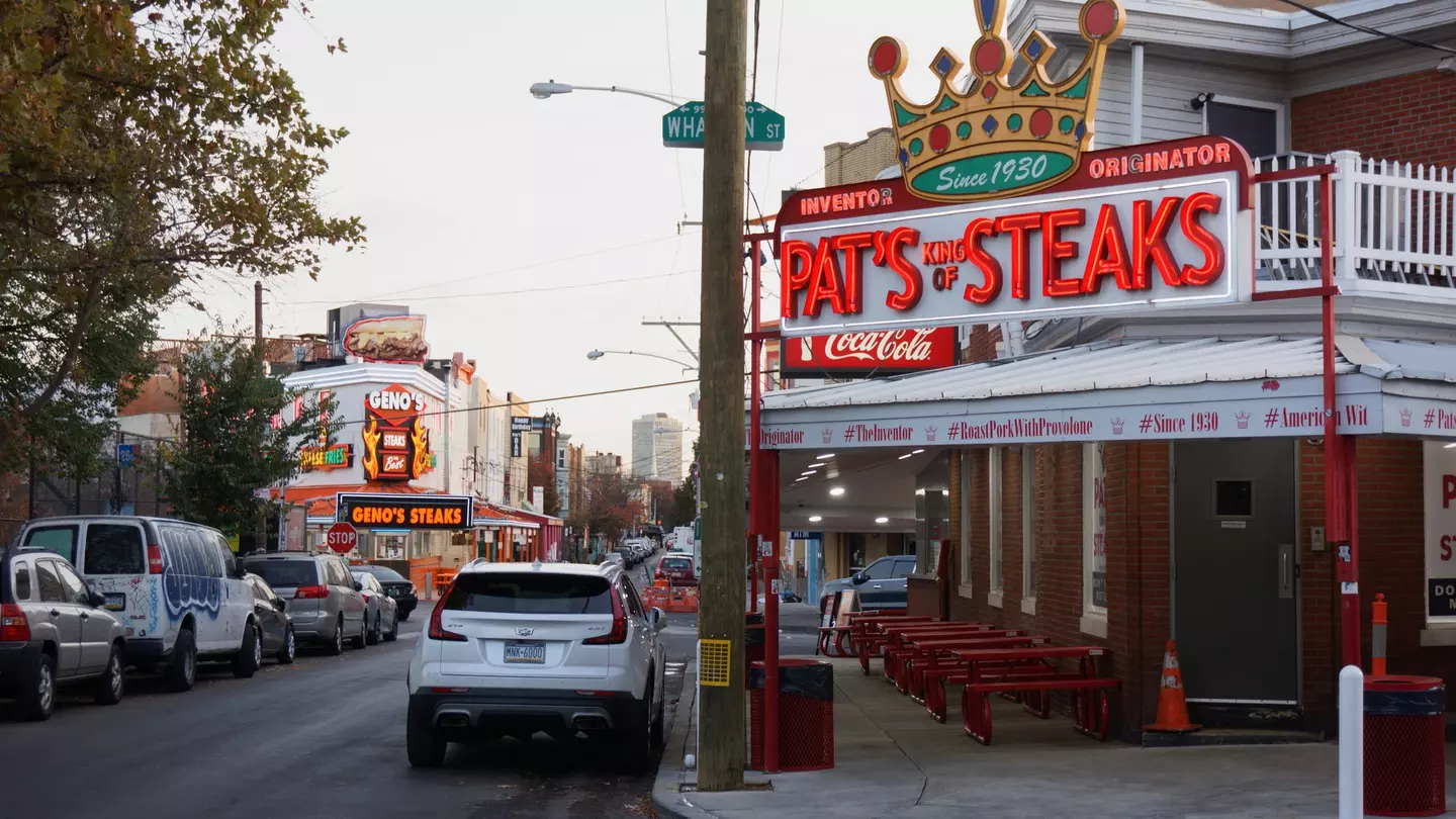 Street lined with cars and a building to the right with a red sign that says "Pat's Steaks".