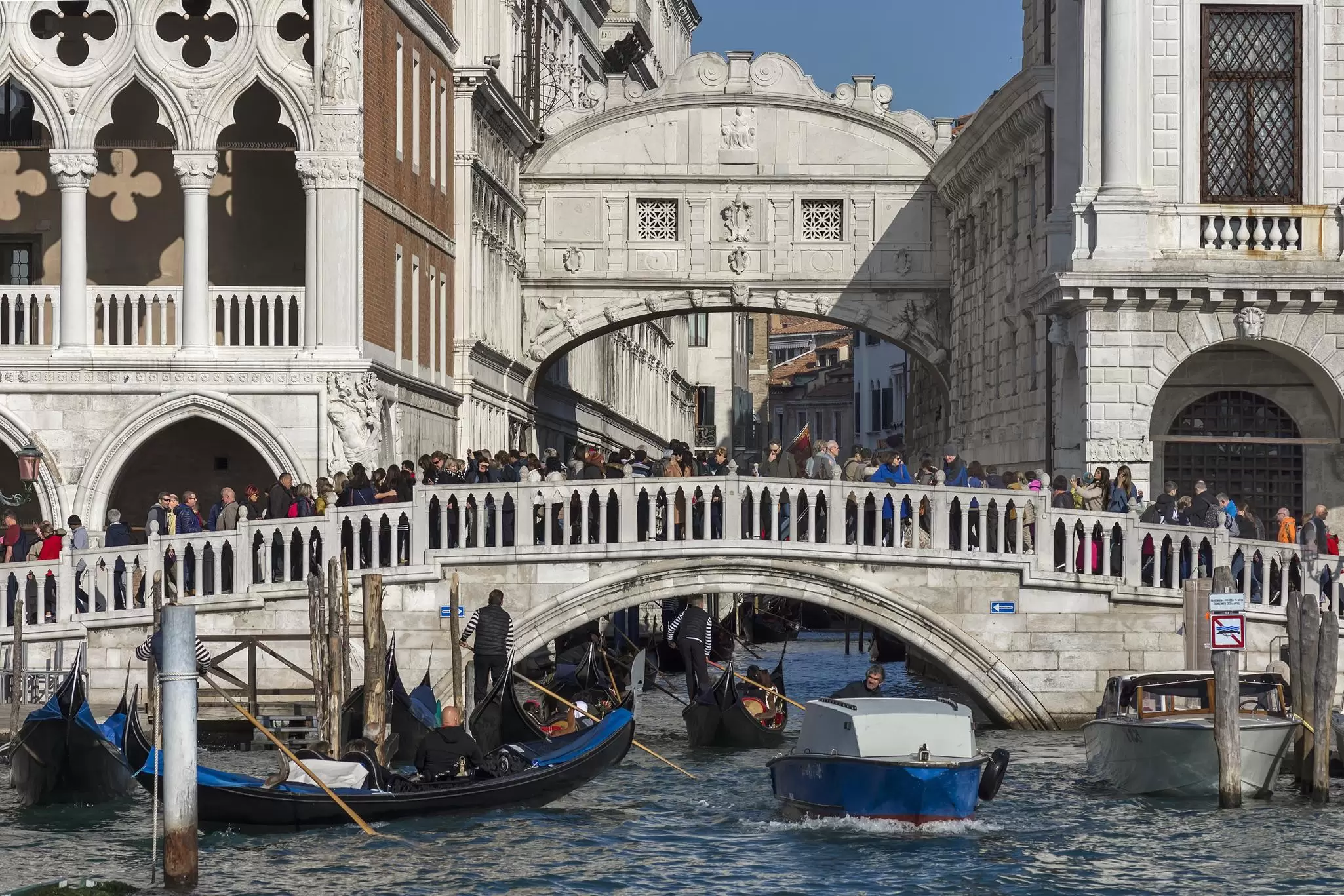Crowds of people are seen on an old stone bridge across a canal in a historic city