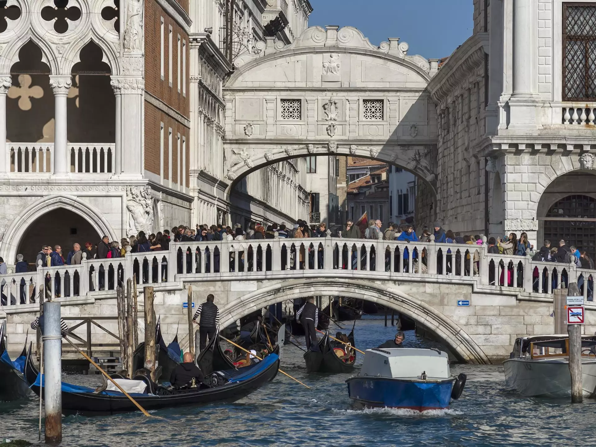 Crowds of people are seen on an old stone bridge across a canal in a historic city