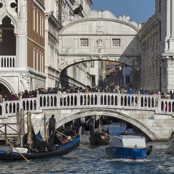 Crowds of people are seen on an old stone bridge across a canal in a historic city