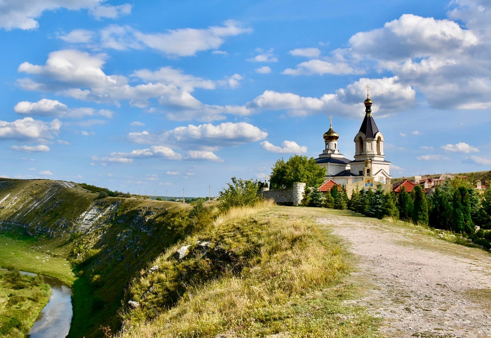 A white church with a gold onion dome and a dark spire standing at the edge of a limestone bluff, above a track and a river winding through the valley below.