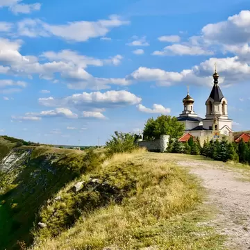 A white church with a gold onion dome and a dark spire standing at the edge of a limestone bluff, above a track and a river winding through the valley below.