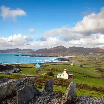 Scattered white and blue houses among green fields beside a turquoise bay, with brown hills under white clouds behind.