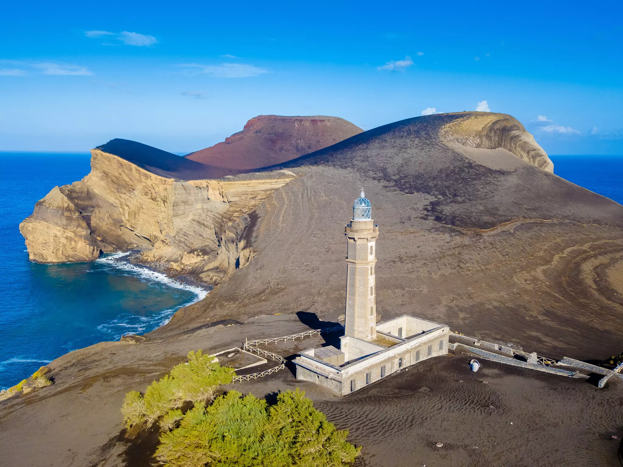 A lighthouse stands at the center of an ash-strewn landscape bordered by bright blue ocean.
