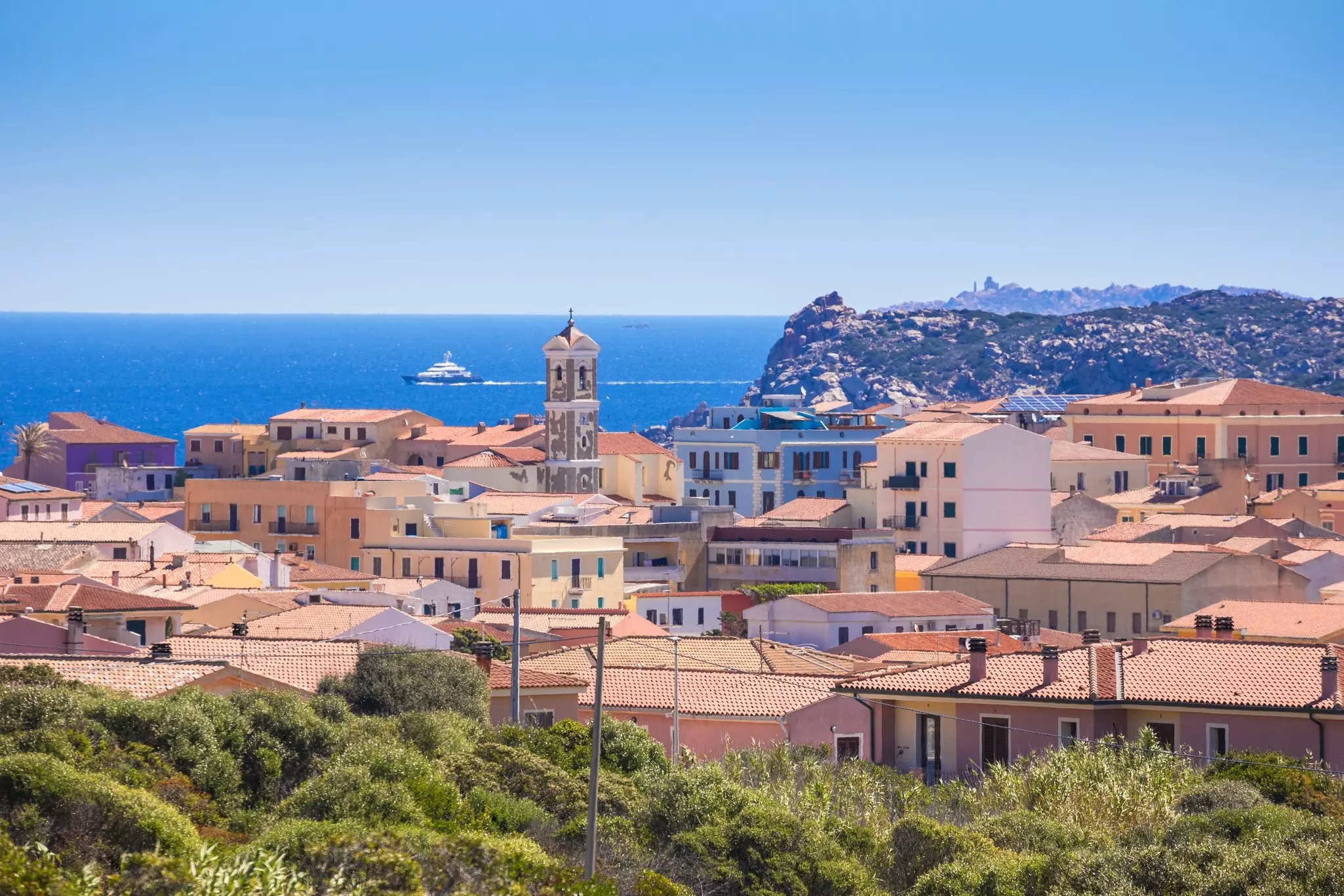 A town in Sardinia set against the blue sea; there are terra cotta roofs on low buildings and a tall tower in the center.
