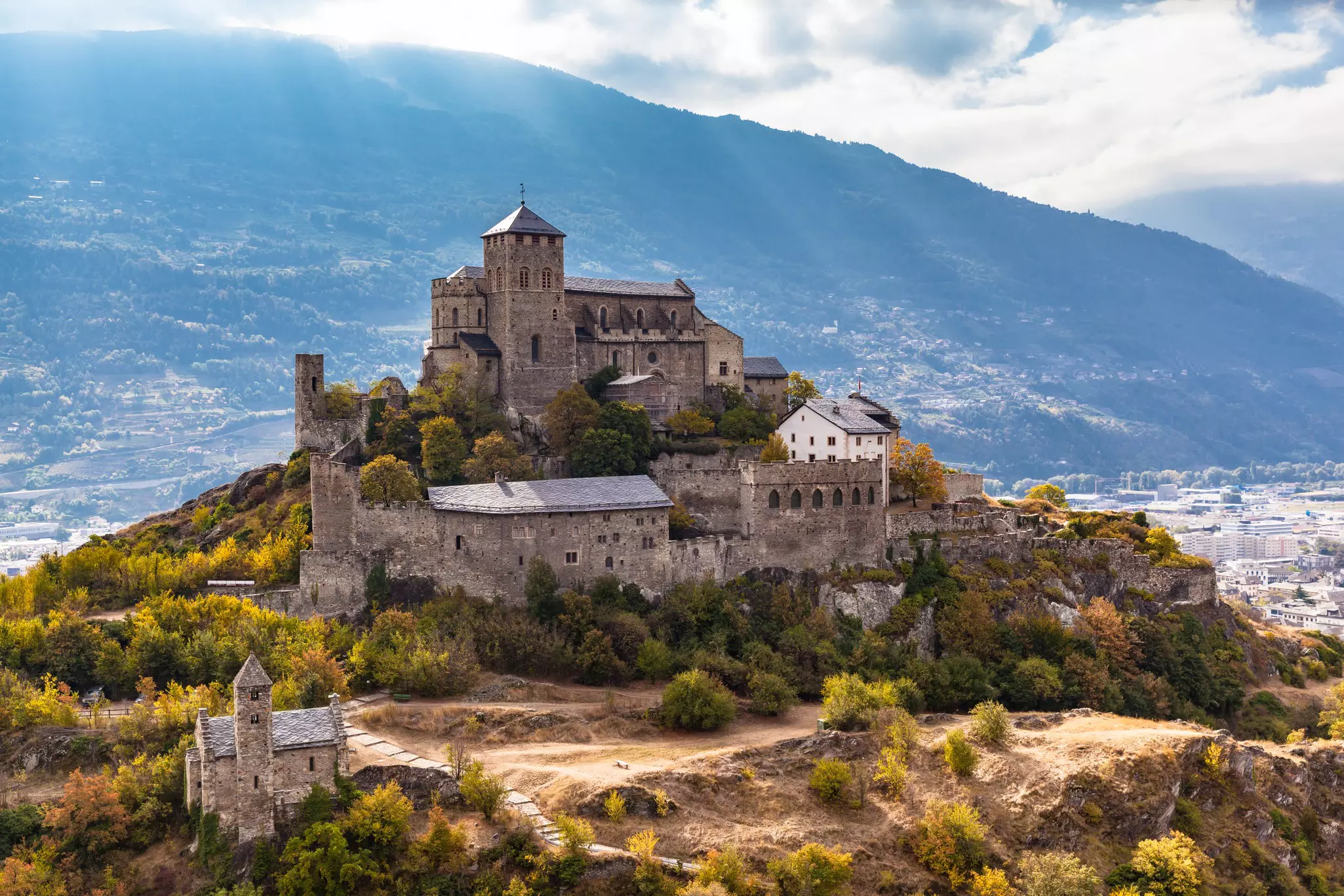 Aerial view of fortified stone church atop a hillside with a forested mountain in the distance and a slight view of a town below on a partly sunny day.