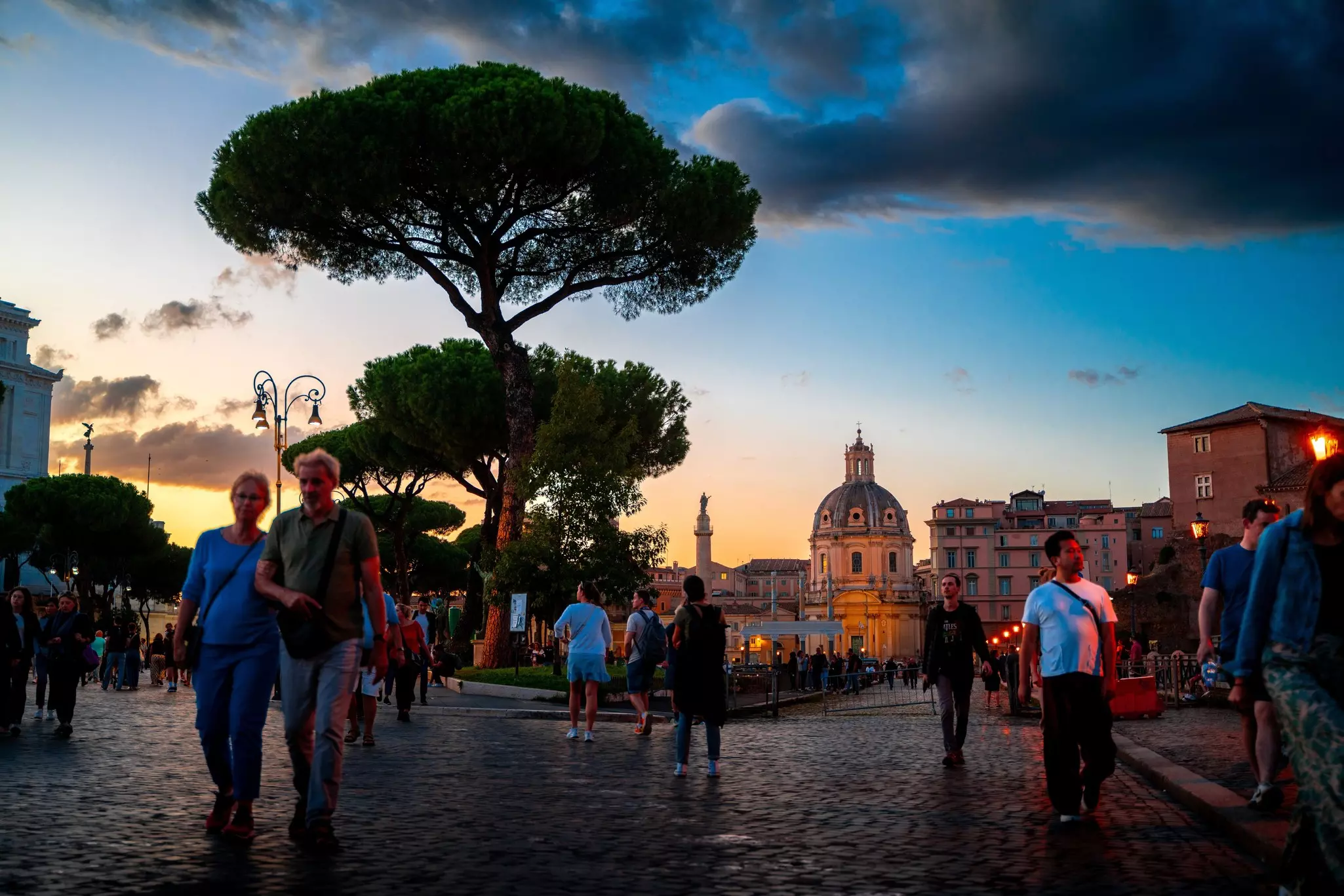 People walk in a city square at dusk, with trees silhouetted and the facade of a church illuminated