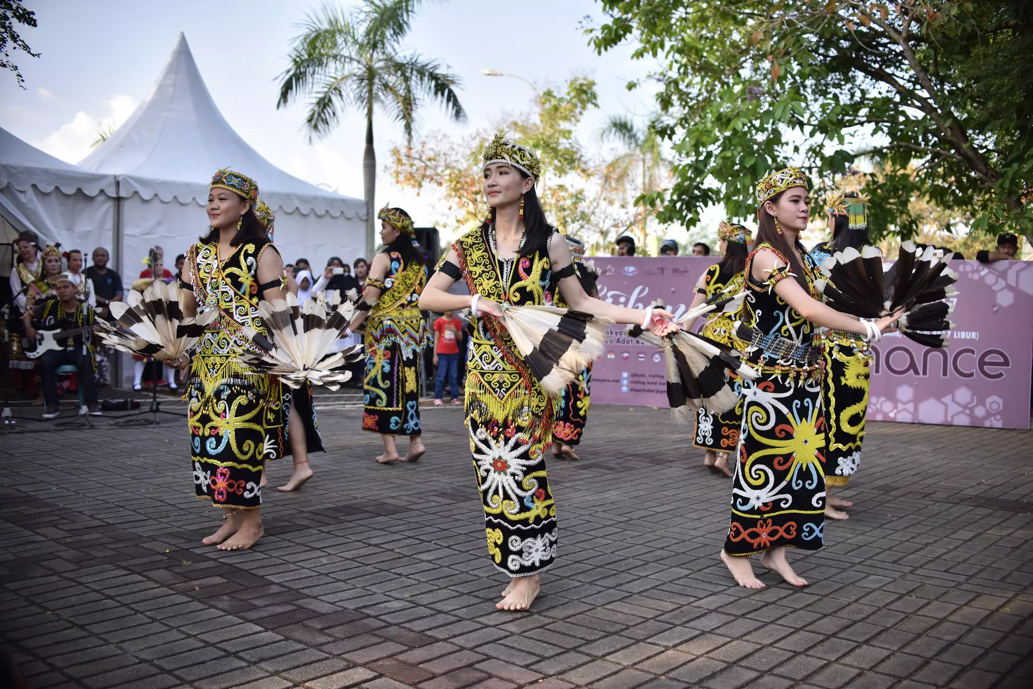 Women in brightly printed dresses wave fans as part of a dance performance at a public festival.