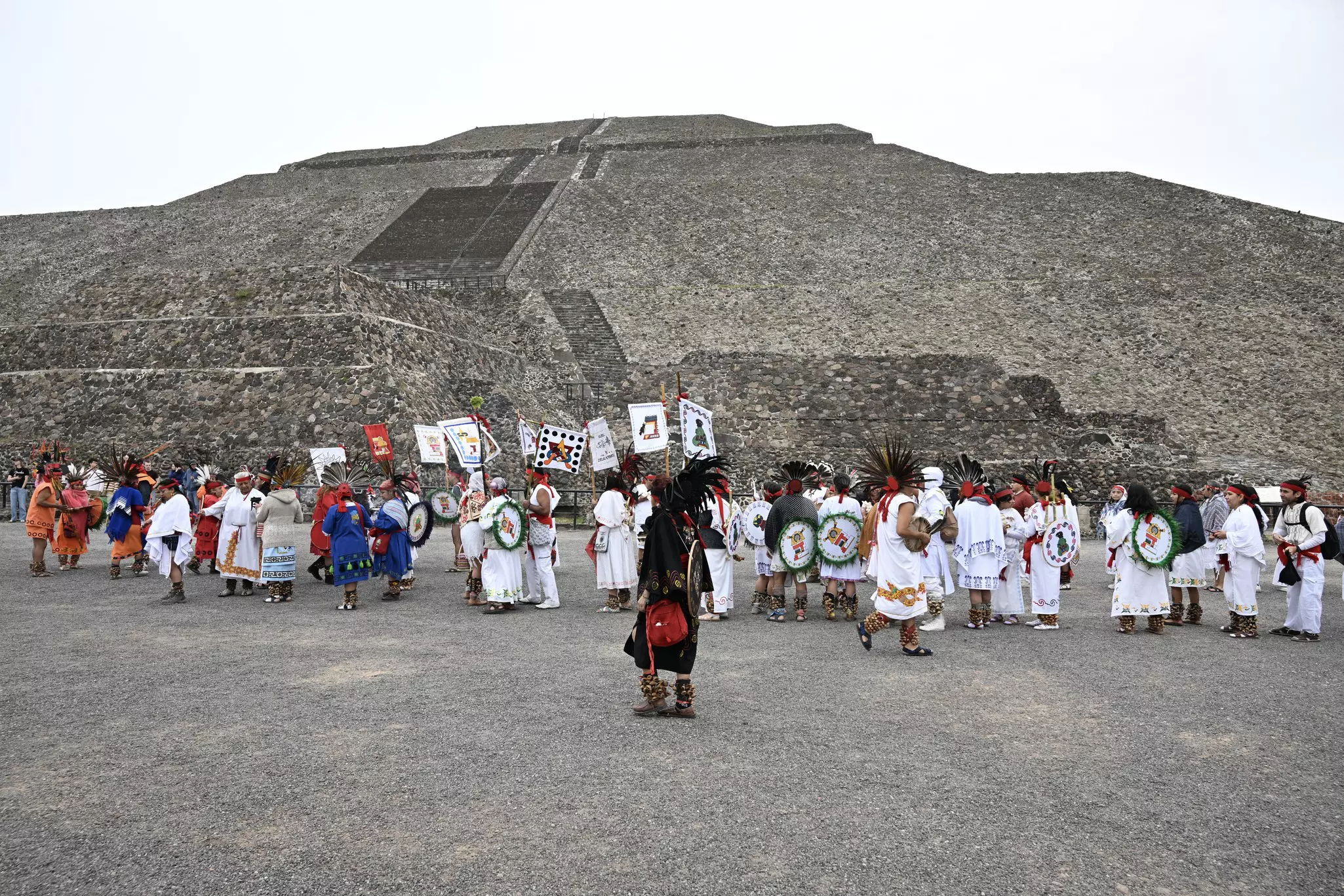 Wide shot of a long line of people in white dress, many with feathered headware, and one man in front with a long black dress in front of a pyramid towering behind them. Many people hold signs.