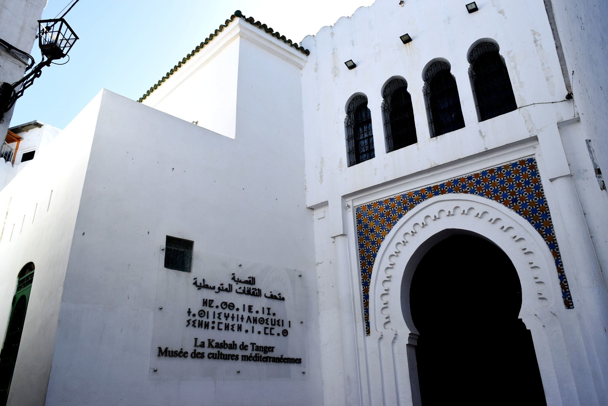 Whitewashed building with a tiled horseshoe archway and a trilingual museum sign.