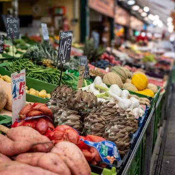A close up shot of a stall at a street market with a white gazebo roof. The stall is selling basil, cheese and crusty bread. A woman in a woolly jumper and brown apron is reaching over to serve a customer.