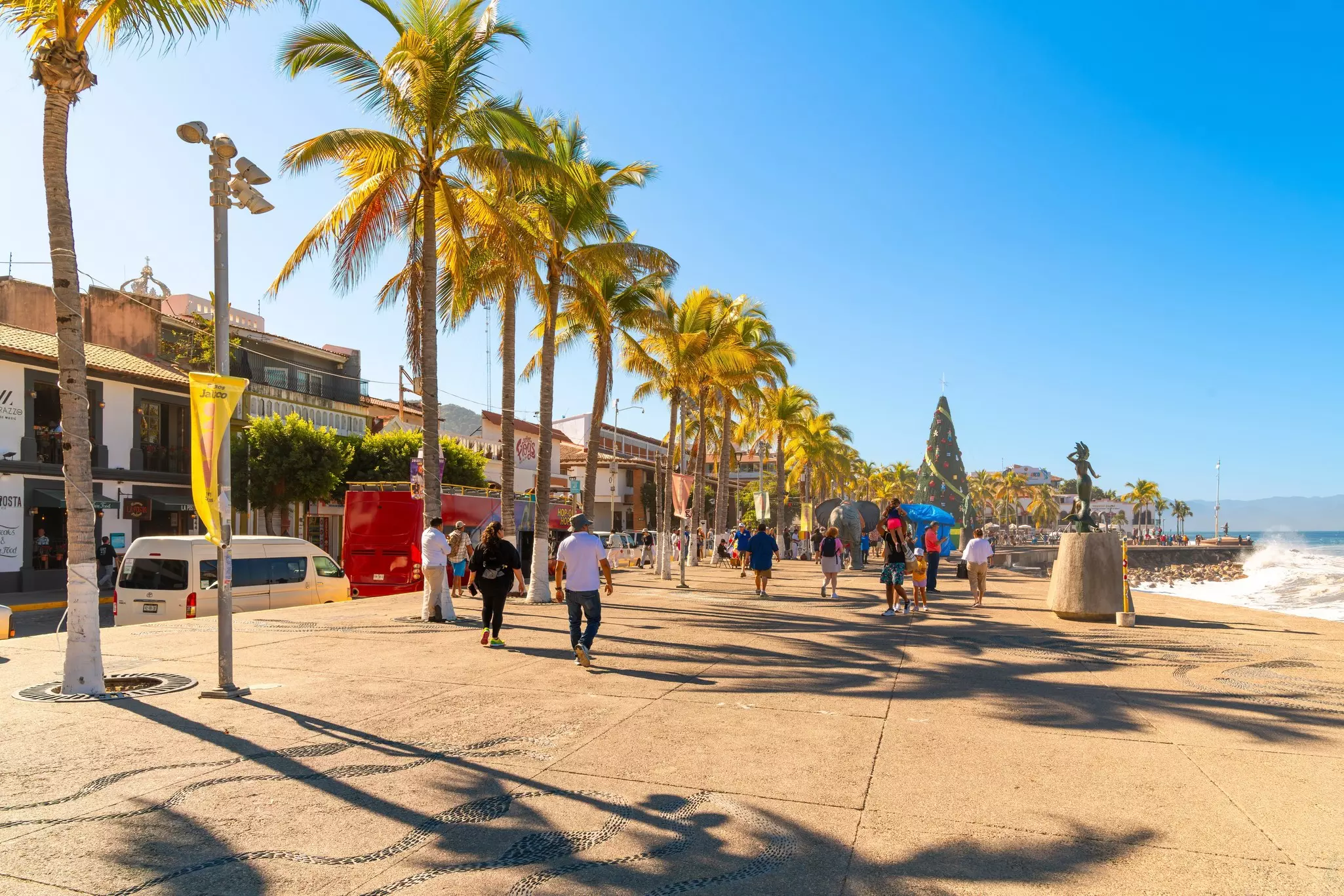 People walk on a wide promenade by the ocean under palm trees.