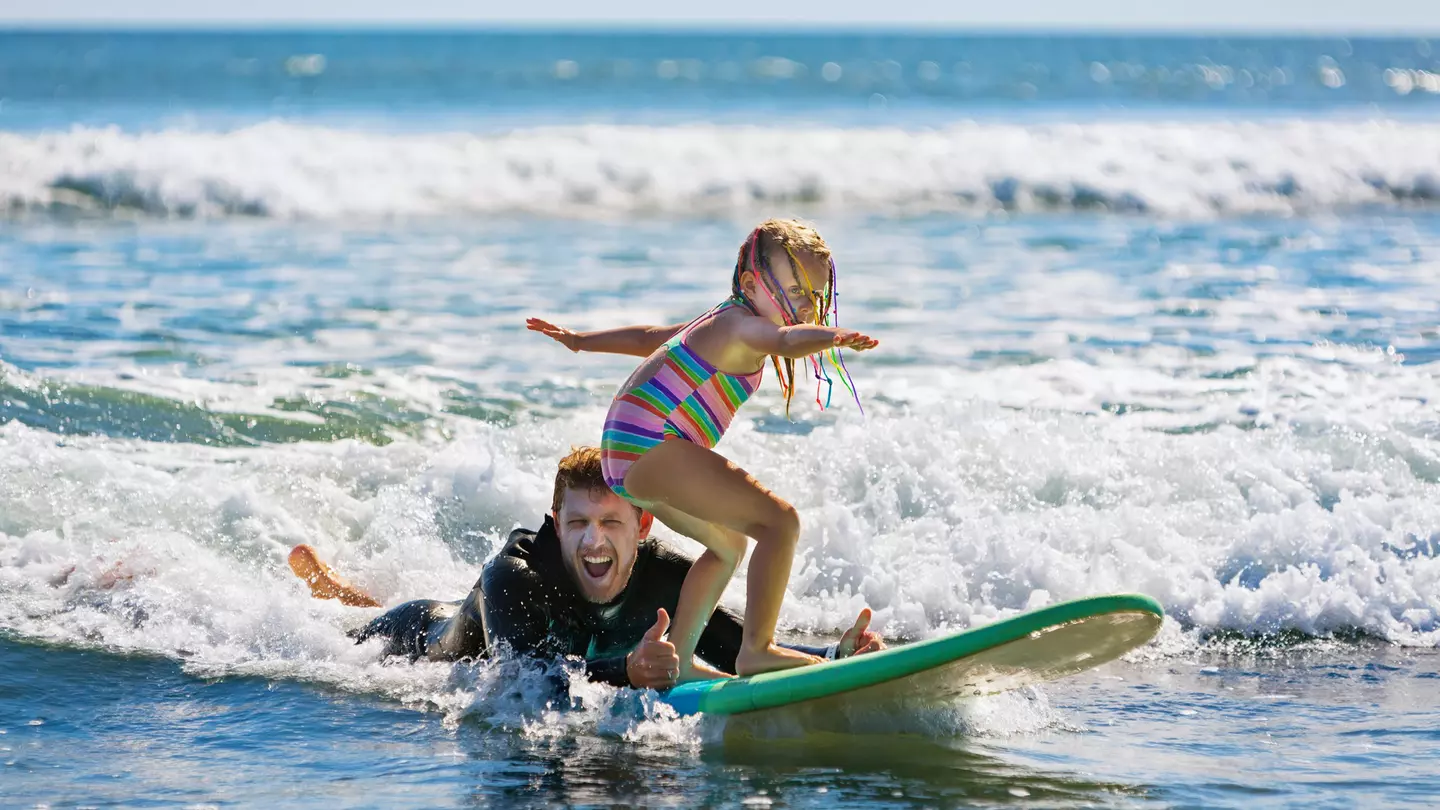 A man in a wet suit lies in the surf giving a thumbs-up signal as a young child balances on a surf board