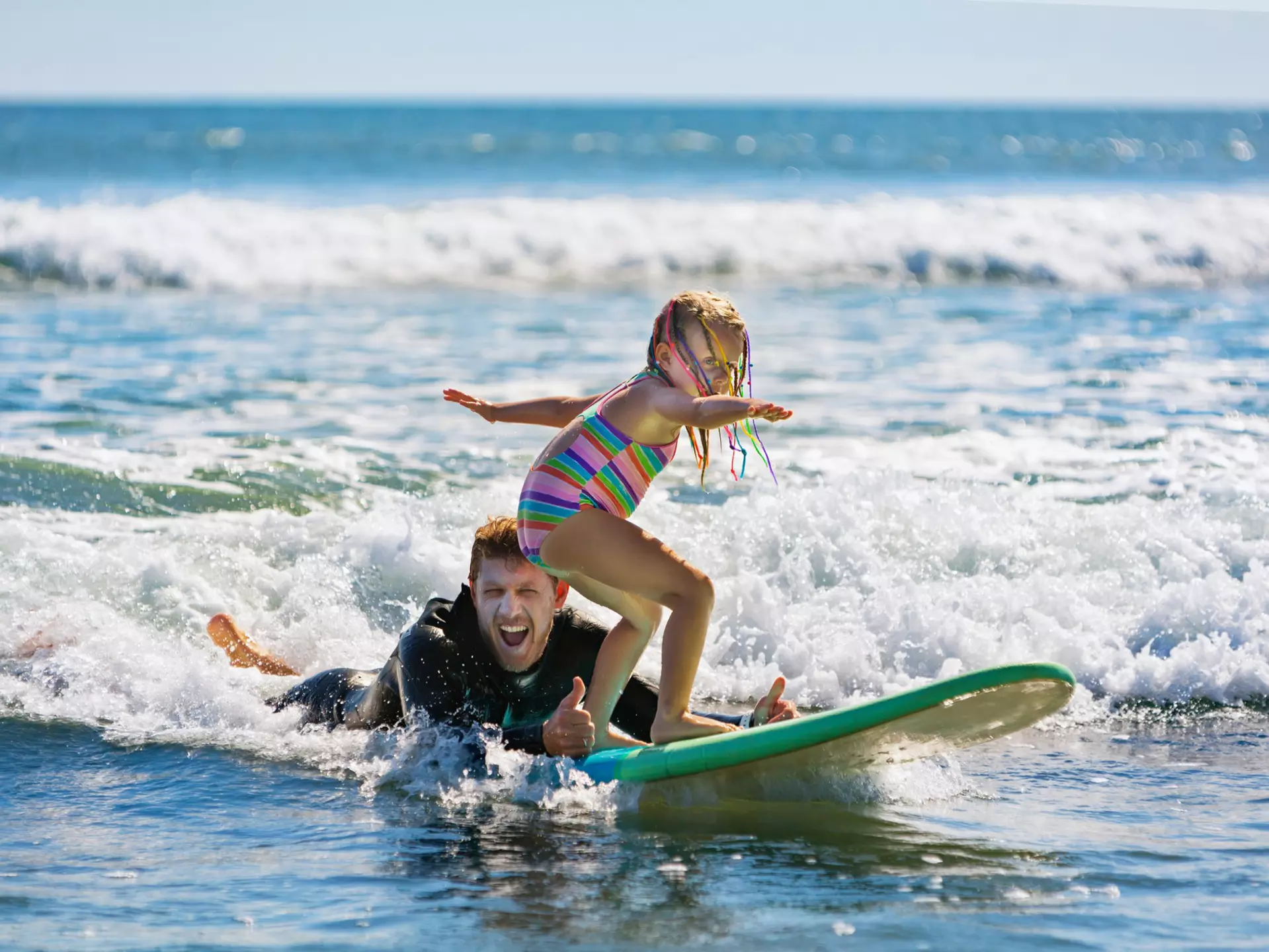 A man in a wet suit lies in the surf giving a thumbs-up signal as a young child balances on a surf board