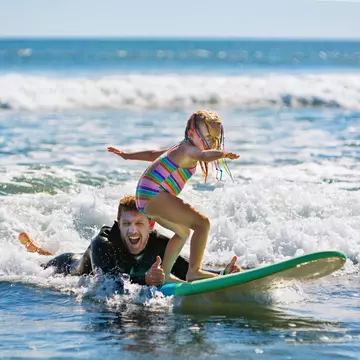 A man in a wet suit lies in the surf giving a thumbs-up signal as a young child balances on a surf board