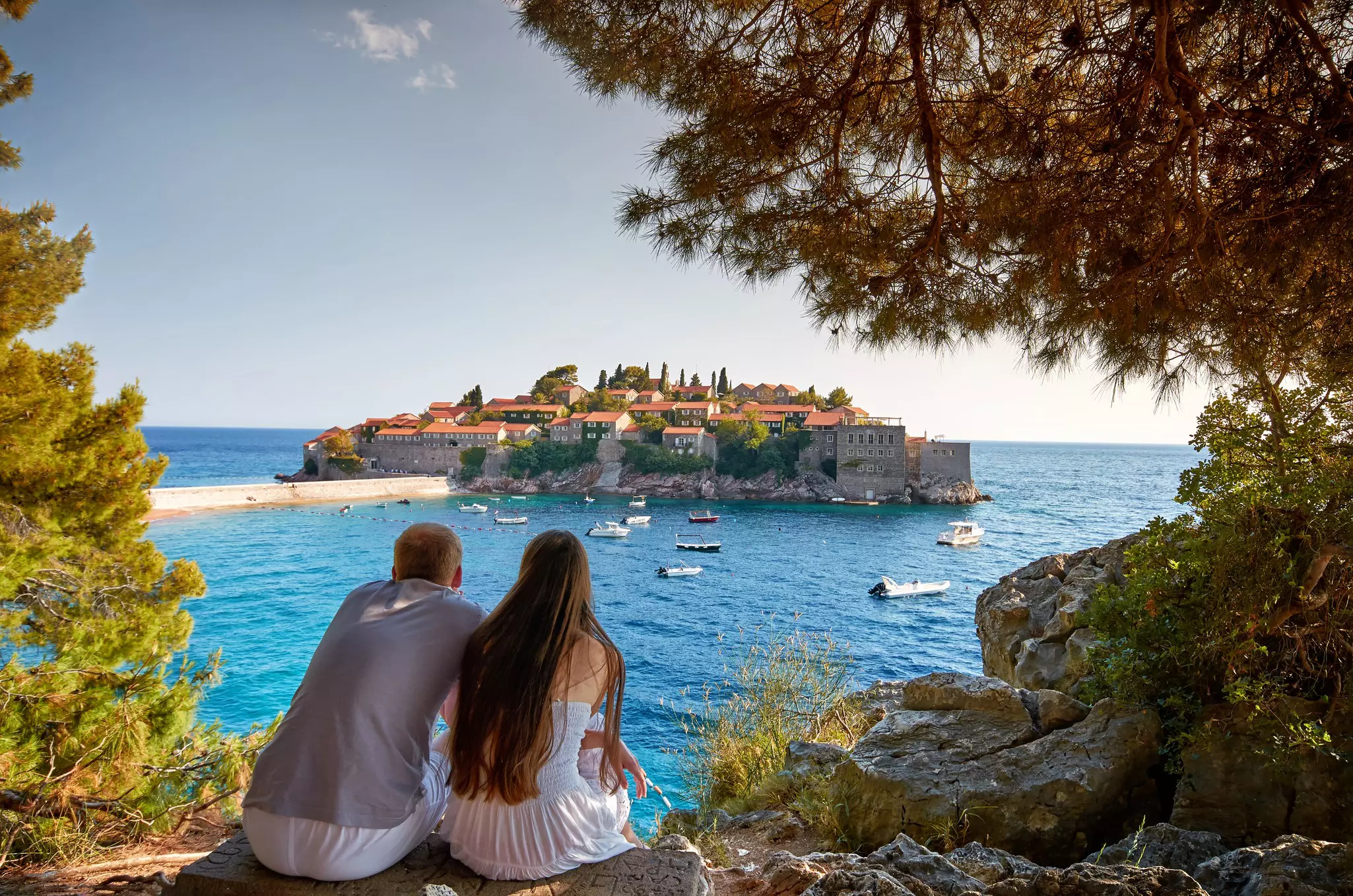 Two people sitting on rocks above a cliff, looking across blue water to an island with low buildings.