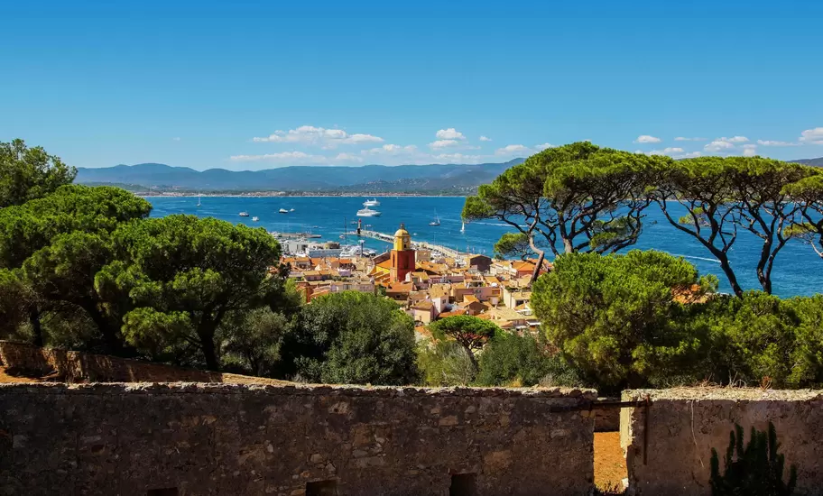 St-Tropez from the citadel: old-town rooftops and an ochre bell tower by the harbour, framed by umbrella pines and blue gulf