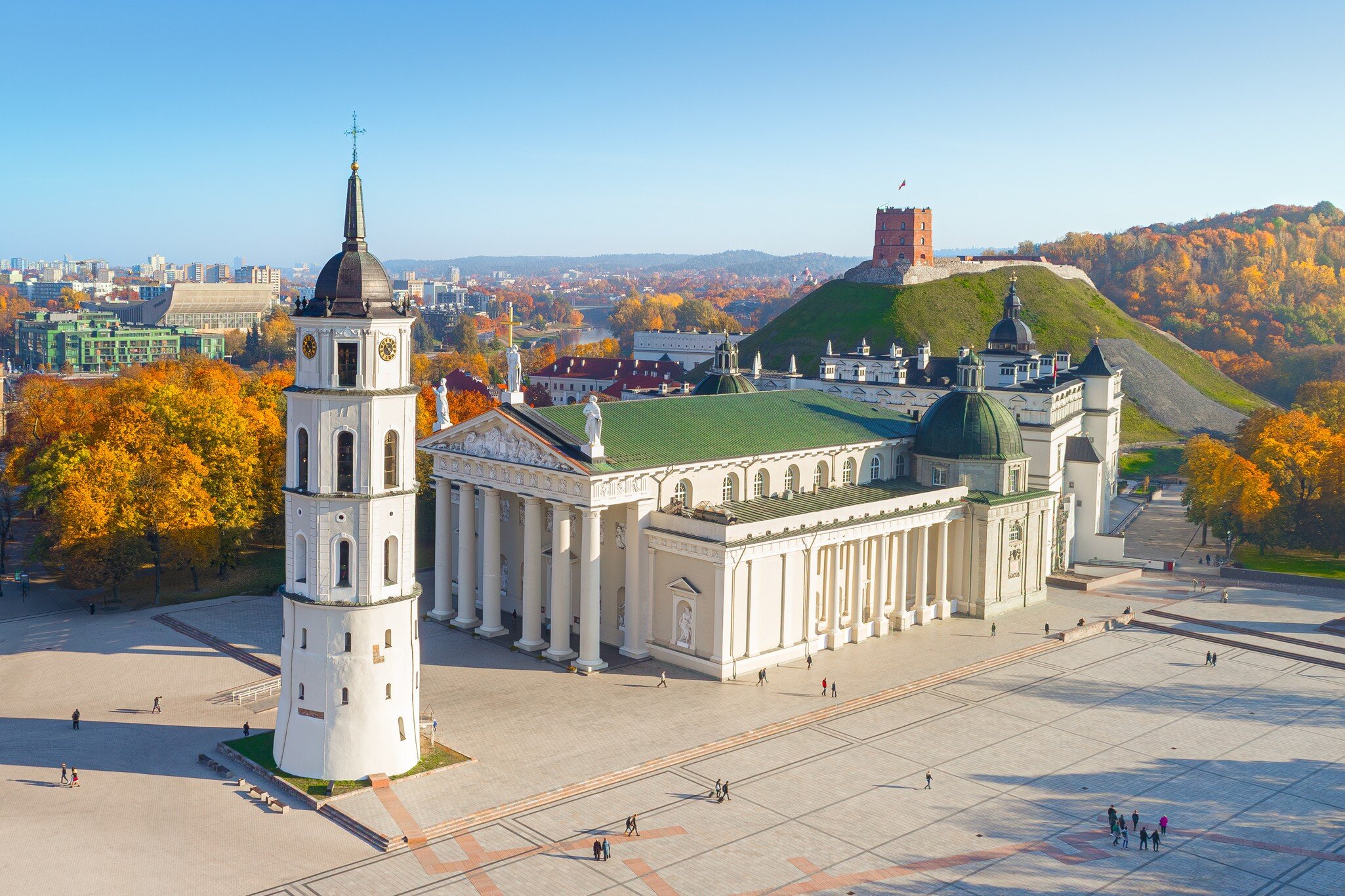 Columned white church with a green roof beside a white bell tower, a red-brick tower on a green hill behind