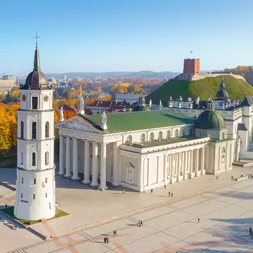 Columned white church with a green roof beside a white bell tower, a red-brick tower on a green hill behind