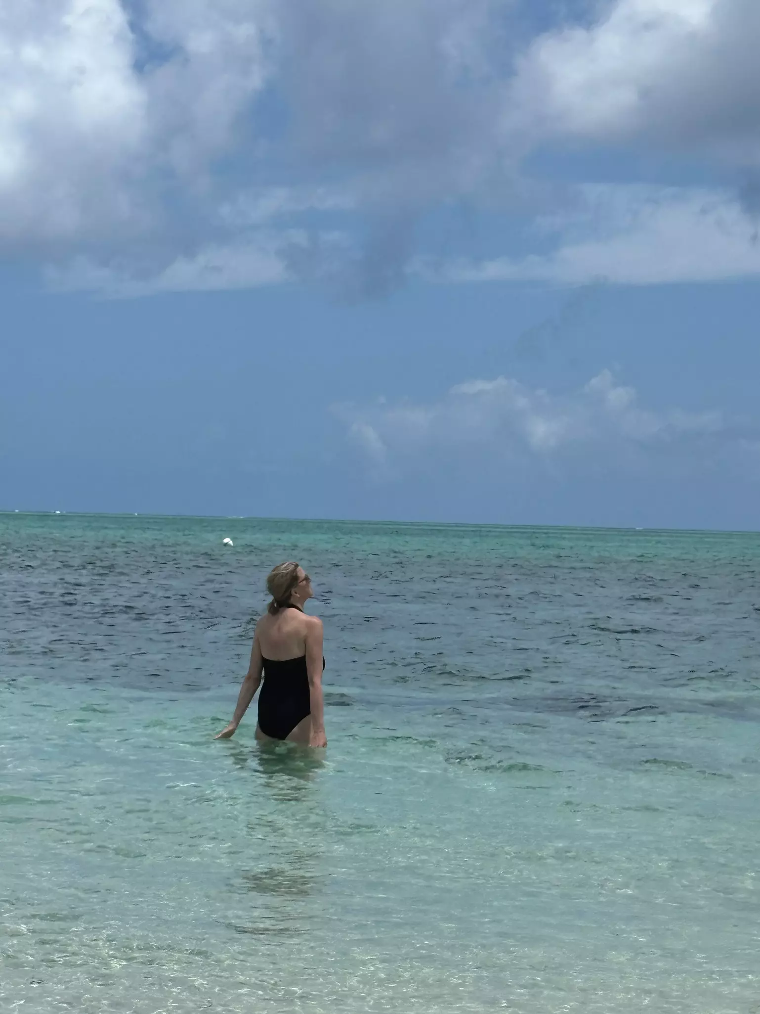 A woman in a black bathing suit wades into the clear turquoise ocean.