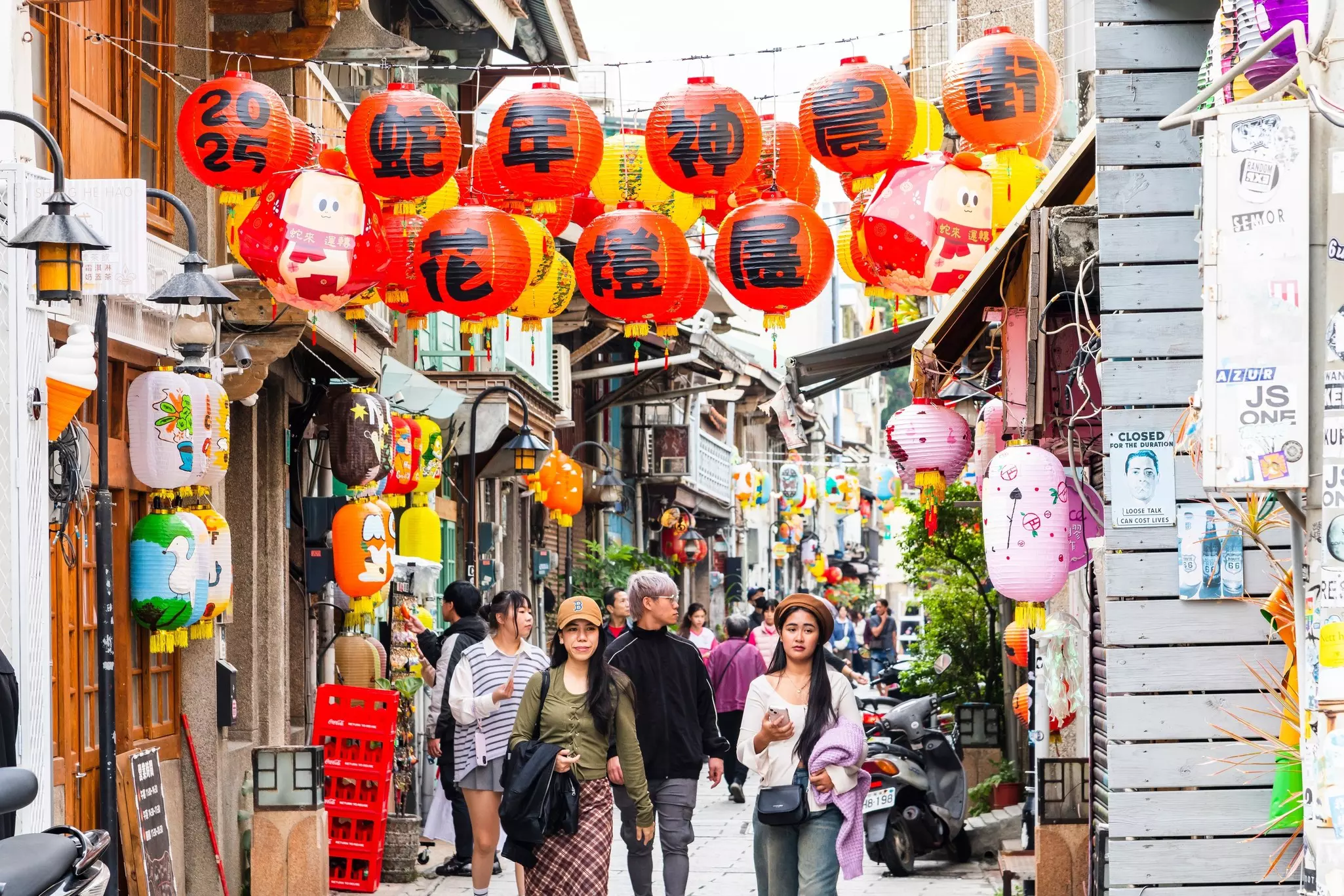 People walk down a narrow lane in a city, under paper lanterns displaying Chinese characters.
